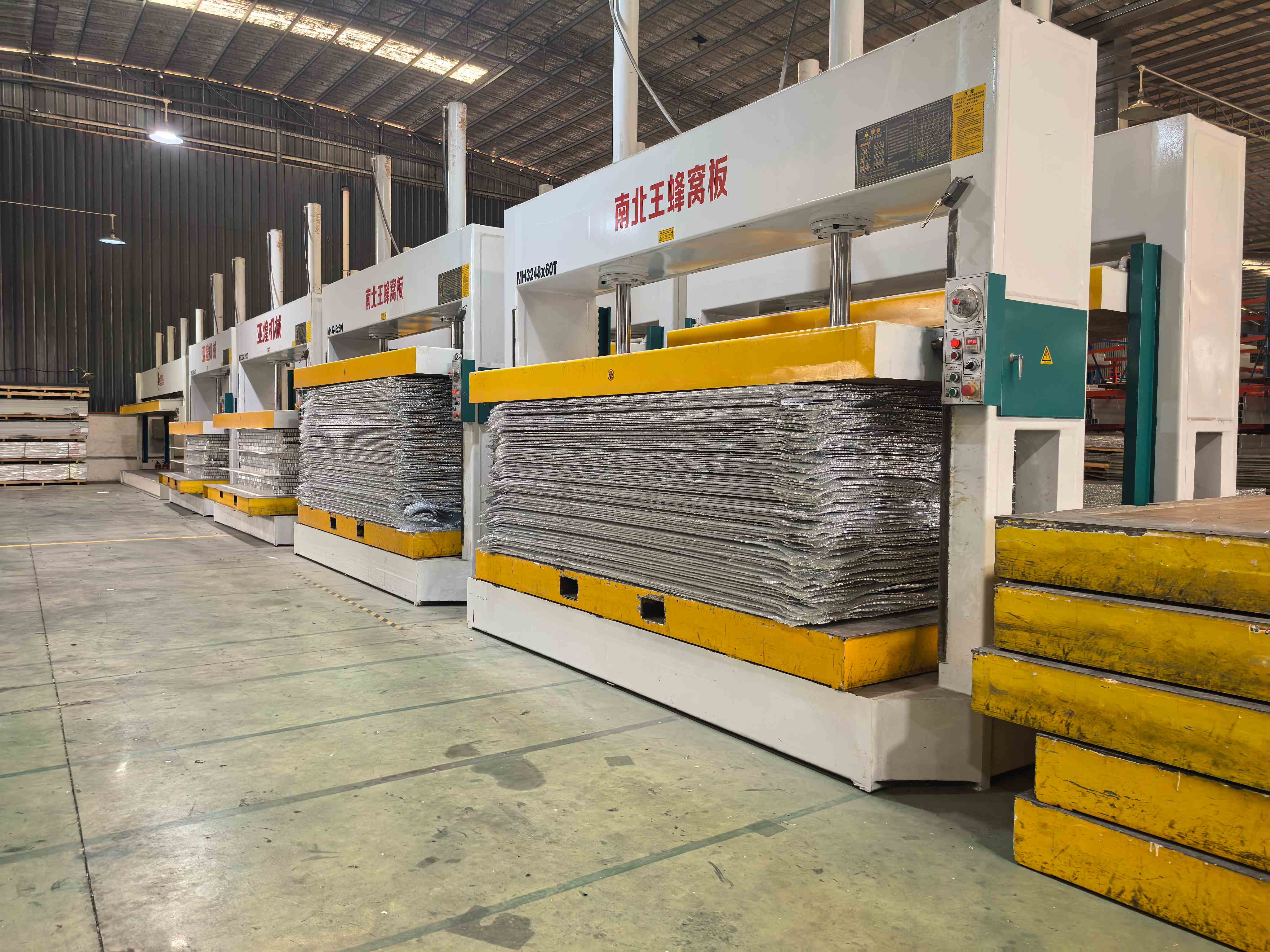 Row of large press machines with stacked reflective panel sheets inside a workshop